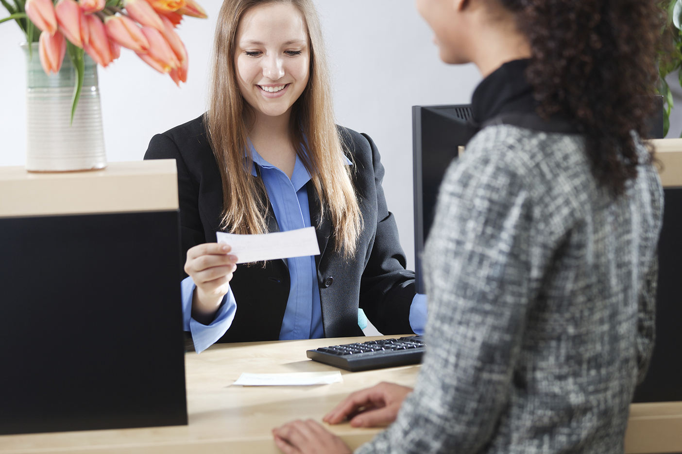  A female bank teller with long brown hair wearing a blue blouse and black suit jacket is assisting a customer with a check.