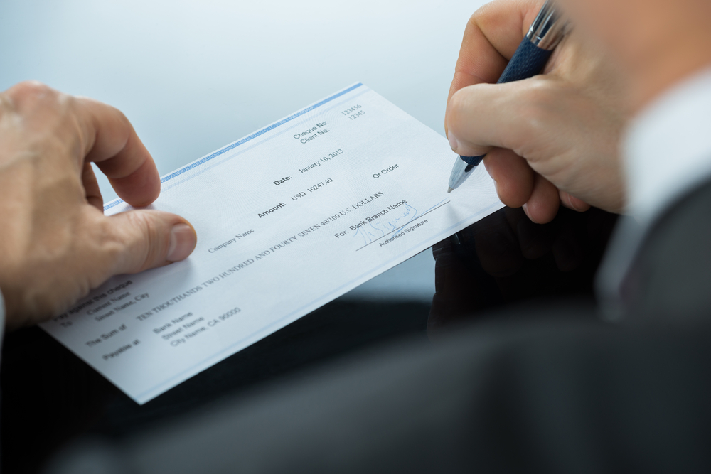 Close-up,Of,Businessman,Hand,Filling,Blank,Cheque,At,Desk